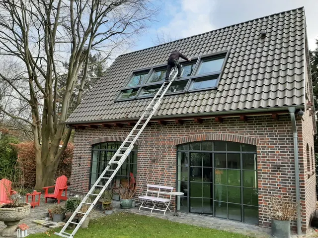 Fensterputzer auf der Leiter reinigt Dachfenster an einem Klinkerhaus